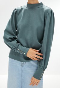 Sweatshirt à col montant bleu sarcelle avec manches longues froncées et boutons aux poignets, associé à une jupe en denim bleu clair.