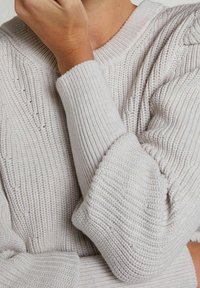 Person som bär en ljusgrå ribbad stickad tröja med uppkavlade ärmar, böjd arm och hand nära ansiktet.