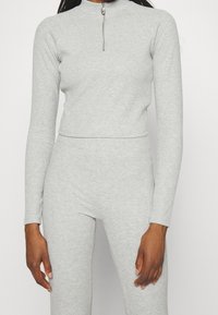 Donna che indossa un top corto a maniche lunghe grigio chiaro a coste con zip frontale e pantaloni aderenti coordinati, su uno sfondo semplice.