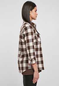 Camicia a quadri marrone e bianca con maniche arrotolate e orlo curvo, realizzata in un tessuto leggero. Presenta una tasca sul petto.