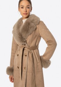 Abrigo de ante sintético beige con un lujoso cuello de piel, cierre de botones doble, y cinturón en la cintura; acentos de piel en los puños.