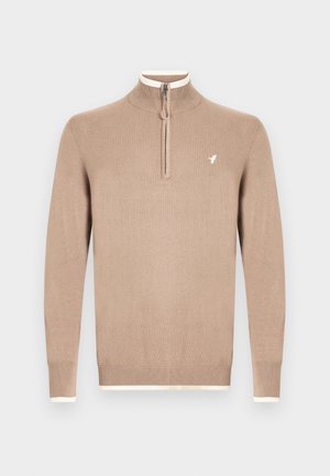 Beige strikket genser med høy krage og halv glidelås. Har kremfarget kant ved kragen og mansjettene, med en liten logo på brystet.