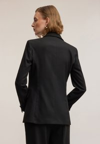 Zwarte op maat gemaakte blazer met gestructureerde schouders, enkele achterventilatie en gladde stof. Gestyled met een klassieke reverskraag en met knopen afgewerkte manchetten.