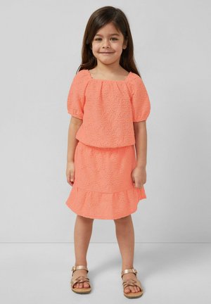 Młoda dziewczyna z długimi ciemnymi włosami, ubrana w koralowy top w wzory z krótkim rękawem oraz pasującą spódnicę i złote sandały, stoi na neutralnym tle.