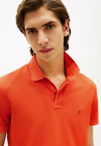 Pomarańczowa koszulka polo z krótkim rękawem, wykonana z fakturowanej tkaniny, z klasycznym kołnierzykiem i małym wyszywanym logo na piersi.