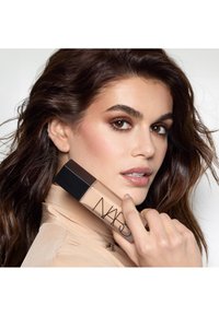 Femme aux cheveux bruns et maquillage tenant une bouteille de fond de teint NARS près de son visage, portant un haut beige sur un fond uni.