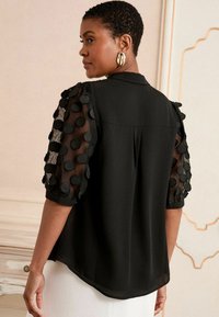 Čierna blúzka so sieťovými balónovými rukávmi zdobená textúrovanými čiernymi bodkami. Prispôsobený strih, zaoblený lem a viditeľný detail gombíka na zadnej strane.