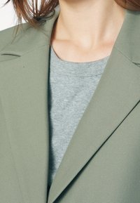 Blazer verde oliva con tessuto testurizzato, caratterizzato da rever a notched e design strutturato, abbinato a un maglione a collo rotondo a costine grigio chiaro.