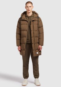 Bruine gewatteerde jas met een capuchon, met een rits en knoopsluiting, gedragen over een olijfgroene hoodie en bijpassende sweatpants. Witte sneakers.
