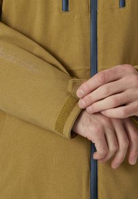 Giacca di lunghezza ginocchio in tessuto giallo senape, con chiusura a strappo sui polsini e una cerniera blu a contrasto. Il marchio è visibile sulla manica.