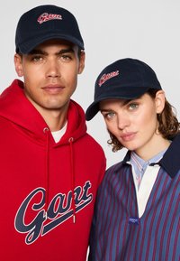 To modeller bruker marineblå baseballcaps med "Gant" i røde sting. Mannen har på seg en rød hettegenser; kvinnen har en blå- og hvitstripete skjorte med krage.