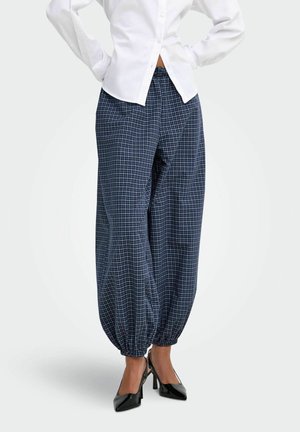 Femme portant un pantalon ample à carreaux bleu marine avec des poignets élastiques, une chemise blanche boutonnée et des chaussures à talons noires, debout les mains sur les hanches.