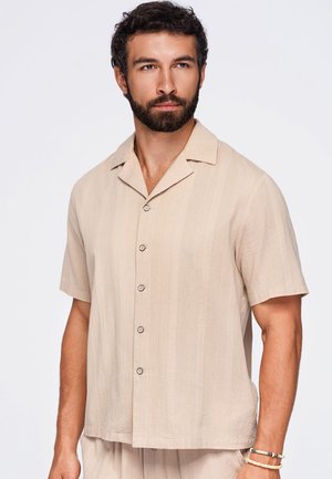 Man met baard en krullend haar, die een beige overhemd met korte mouwen en knopen draagt en armbanden om zijn rechterpols tegen een effen achtergrond.