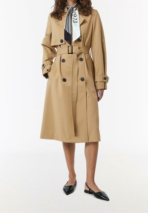 Vrouw gekleed in een beige dubbelgeknoopte trenchcoat met riem, zwarte slingback platte schoenen, en een zwart-wit gestreepte sjaal met de tekst "1973".