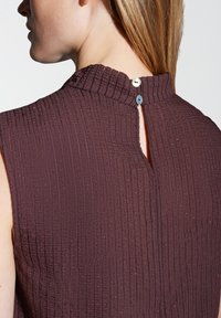 Lila strukturierte Bluse mit vertikalen Streifen, hohem Kragen und einem Schlüsselloch-Design auf der Rückseite, gesichert durch zwei Knöpfe. Leicht glitzernder Stoff.