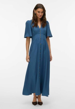 Nő kék, rövid ujjú, gombos maxi ruhában, testhezálló derékkal és barna, hegyes orrú lapos cipőben, fehér háttér előtt áll.