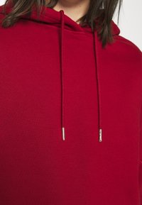 Gros plan d'une personne portant un sweat à capuche rouge uni avec cordons et embouts métalliques, montrant la partie supérieure de la poitrine et le cou.