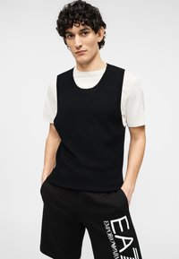 Μαύρο πλεκτό αμάνικο γιλέκο με στρογγυλή λαιμόκοψη, φορεμένο πάνω από άσπρο πουκάμισο, συνδυασμένο με μαύρο σορτς που φέρουν λευκό τυπωμένο λογότυπο.