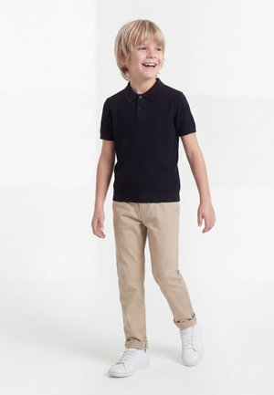 Plavokosi dječak koji se smije, hoda, nosi crnu polo majicu kratkih rukava, bež hlače s manšetama i bijele tenisice, na jednobijeloj pozadini.