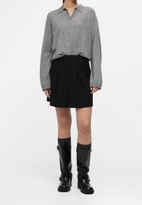 Osoba ubrana w szary sweter z długim rękawem, czarną plisowaną spódnicę, szare skarpetki i czarne kozaki za kolano stoi na białym tle.