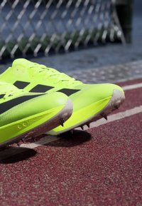 Des pointes de course jaune vif à rayures noires reposant sur une piste rouge près d'une clôture en treillis métallique.