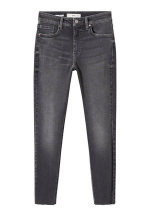 Person iført en løs hvid skjorte med knapper og opkørte ærmer, stoppet ned i højtaljede mørkegrå jeans, hænderne bag ryggen.