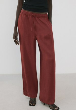 Mujer con pantalones de talle alto y corte amplio de color óxido con cintura de cordón, sandalias negras con tacón y top sin mangas negro.