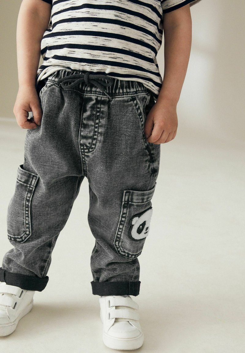 Enfant portant un t-shirt rayé noir et blanc, un pantalon en jean gris avec un patch panda et des baskets blanches, debout sur un sol clair.