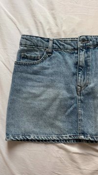 Denim mini krilo v svetlo modri barvi, z sprednjimi žepi, zadrgo in gumbom za zapiranje ter raztrganim robom. Gladka tekstura.