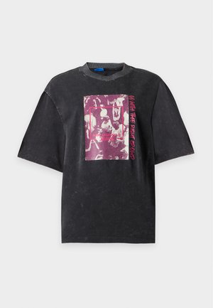 Femme aux cheveux bouclés portant un t-shirt noir avec un motif graphique et un jean large bleu clair, debout devant un fond uni.