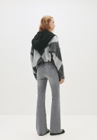 Mladá žena v sivých rozšírených džínsoch, čiernych topánkach a svetri s šedou a čiernou argyle vzorom a čiernou kapucňou, stojaca bokom.