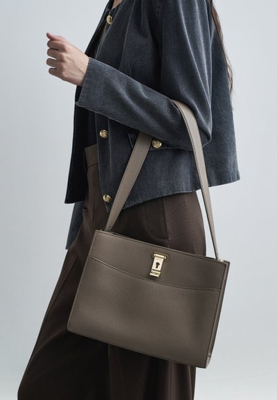 Bežna torbica z teksturo, ploske oblike in zlatim okovjem. Nošena na rami z sivo jakno, ki ima zlate gumbe.