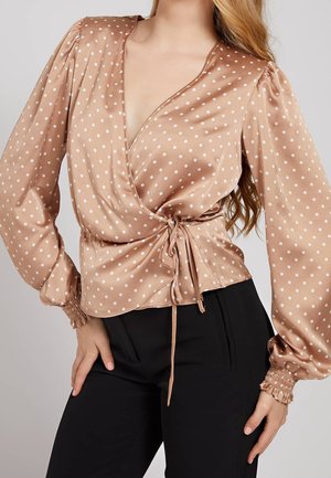 Vrouw draagt een beige satijnen wikkelblouse met witte polkadots en opgeblazen lange mouwen, gestrikt in de taille, gecombineerd met een zwarte broek.