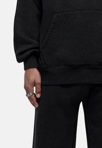 Felpa nera con tasca frontale, polsini e orlo a coste, abbinata a pantaloni della tuta neri coordinati. Mano visibile con anello d'argento.
