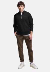 Zwarte rits-hals trui gemaakt van gebreide stof, gecombineerd met bruine corduroypakken en witte sneakers. Voorzien van een cirkelvormig logo op de borst.