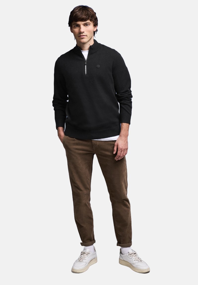 Zwarte rits-hals trui gemaakt van gebreide stof, gecombineerd met bruine corduroypakken en witte sneakers. Voorzien van een cirkelvormig logo op de borst.