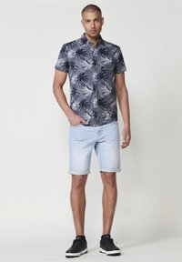Camisa de manga corta azul marino con patrón de hojas de palma, combinada con pantalones cortos de mezclilla claros. Zapatillas negras completan el atuendo.