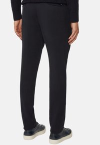 Mies seisoo yllään räätälöidyt mustat housut ja nahkakengät, näyttäen takanäkymän, kädet sivuilla.