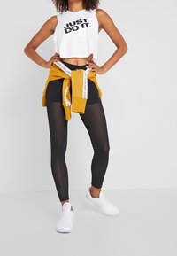 Donna che indossa un top bianco corto "Just Do It", leggings neri, sneakers bianche e una felpa senape legata in vita.