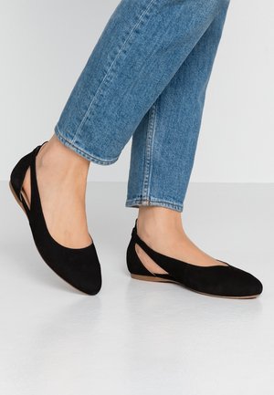 Kvinde iført blå jeans og sorte lukkede slingback-flade sko står på en glat hvid gulv med den ene fod let hævet.