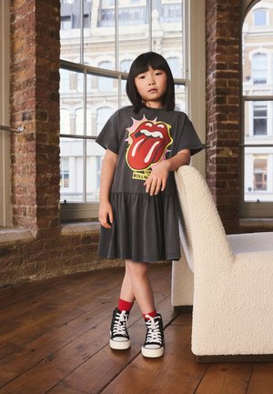Enfant portant une robe noire Rolling Stones avec des chaussettes rouges et des baskets montantes noires, appuyé sur une chaise blanche texturée dans une chambre avec de grandes fenêtres.