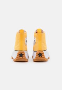 Żółte płócienne botki za kostkę z białą podeszwą, czarną gwiazdą, okrągłą naszywką z logo i teksturowaną podeszwą gumową. Widok z tyłu z kontrastującymi detalami.