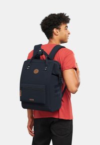 Sac à dos bleu marine avec un design structuré, poignées supérieures, bretelles ajustables, poche zippée à l'avant et un accent de logo rond. Tissu texturé.
