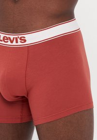 Rde bombažne bokserice z belim pasom, ki nosi rdeči napis "Levi's", elastičen material, prilegajoč dizajn in brezšivne robove.
