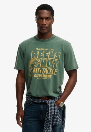 Man draagt een groen T-shirt met de tekst 'Reels Hut Bait & Tackle Superdry', blauwe spijkerbroek, geruit overhemd om de taille gestrikt en een polshorloge.