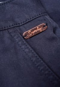 Navy fabric with a copper tag labeled "Superdry." Features stitched edges and smooth texture. Tag attached with black thread.