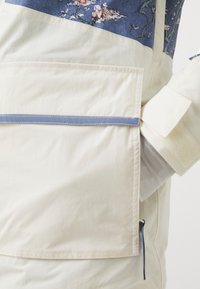 Lagana krem jakna s cvjetnim uzorkom u plavoj gornjoj polovici, džepom na patent zatvarač i podesivim manžetama sa čičak trakama.