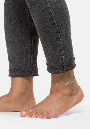 Bracelet de cheville en argent avec un pendentif croix sur un pied nu ; la personne porte un jean skinny grisé foncé replié. Texture de peau et détails des ongles bien visibles.