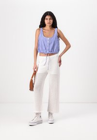 Blau-weißer gestreifter kurzer Tanktop mit Knöpfen, kombiniert mit weißen gestrickten weit geschnittenen Hosen und weißen High-Top-Sneakern. Braune Schultertasche.