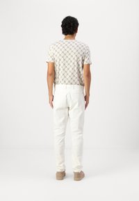Homme debout de dos, portant une chemise à manches courtes à motif, un pantalon blanc et des chaussures en daim beige, sur un fond uni.